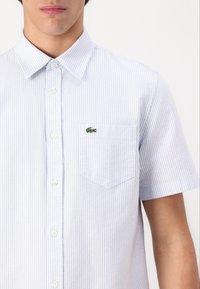 Vaaleansininen lyhythihainen paita, jossa on valkoiset pystysuorat raidat, napit edessä, terävä kaulus ja pieni vihreä krokotiililogo rintataskussa.