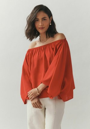 Frau mit schulterlangem dunklem Haar, die eine rote schulterfreie Bluse, weiße Hose, ein goldenes Armband, eine Halskette und Ohrringe vor einem einfarbigen Hintergrund trägt.