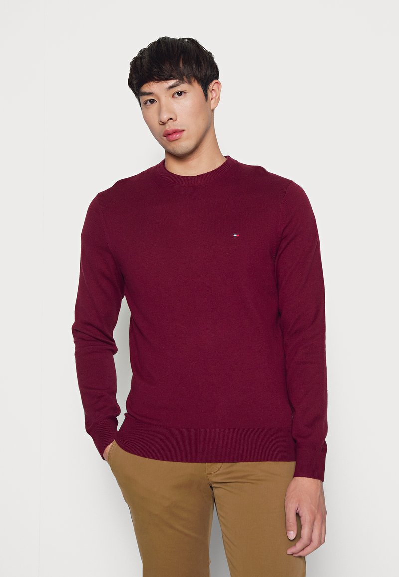 Burgunderfarbener Strickpullover mit Rundhalsausschnitt, gerippten Bündchen und Saum, mit einem kleinen Logo auf der Brust. Kombiniert mit khakifarbenen Hosen.