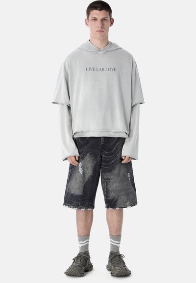 Jeune homme debout portant un sweat à capuche gris surdimensionné avec le texte "LIVE LAB LOVE", un short noir déchiré au genou, des chaussettes grises et des baskets texturées.