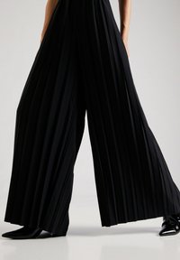 Svarta plisserade byxor med vida ben i mjuk tyg, med hög midja och subtil lyster, kompletterat med spetsiga svarta skor.
