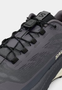 Chaussure de sport grise avec une tige en maille, un motif texturé, des lacets noirs et un embout renforcé. Logo de marque notable sur le côté.