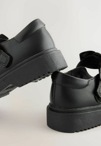 Zapatos sintéticos negros con un acabado texturizado, que cuentan con una suela gruesa, correa ajustable y un diseño minimalista con costuras sutiles.