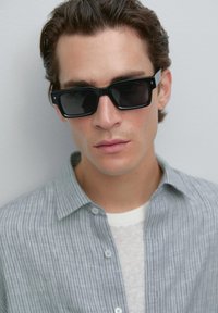 Young man wearing rectangular black sunglasses, gray striped button-up shirt over white top, with short, styled brown hair against plain background.