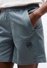 Άτομο που φοράει γκρι σορτς με ελαστική μέση και πλαϊνές τσέπες, με λογότυπο μαμούθ, το χέρι να στηρίζεται μέσα σε μία από τις τσέπες.