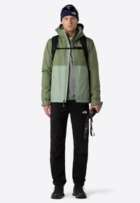 Vihreä ja vaaleanvihreä vedenpitävä takki, harmaa paita, mustat housut, musta asuste kädessä, mustat saappaat ja sininen pipo. Reppu mukana.