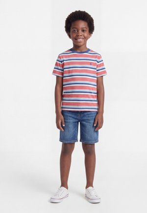 Αγόρι που στέκεται και χαμογελάει, φοράει μια κόκκινη, λευκή και μπλε ριγέ μπλούζα, τζιν σορτς και άσπρα αθλητικά παπούτσια, σε απλό λευκό φόντο.