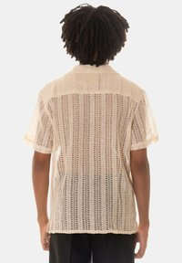 Camicia beige a maniche corte realizzata in un tessuto knit leggero e trasparente con un motivo a righe verticali e un colletto standard.