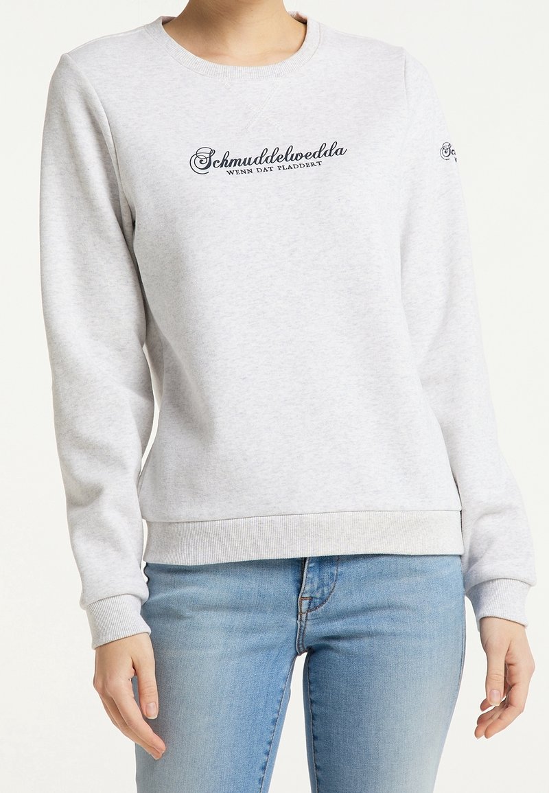 Sweat-shirt gris clair avec texte en cursive noire "Schmuddelwedda wenn dat pladdert" porté avec un jean bleu clair, visible du cou aux hanches.