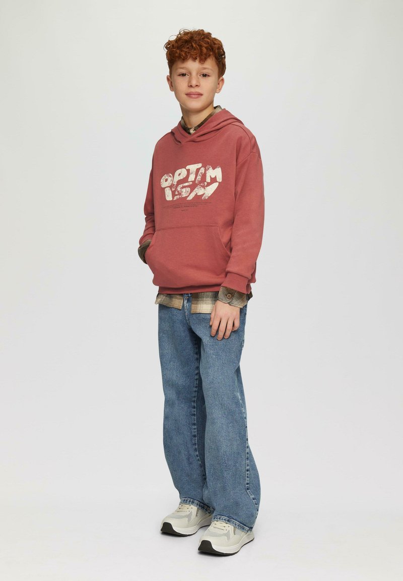 Roze hoodie met witte grafische tekst, blauwe spijkerbroek en grijze sneakers. Hoodie heeft een kangoeroezak en een verstelbare trekkoordkap.