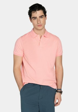 Joven con cabello rizado oscuro, vestido con una camiseta polo rosa claro y pantalones azul oscuro, de pie con una mano en el bolsillo contra un fondo blanco.