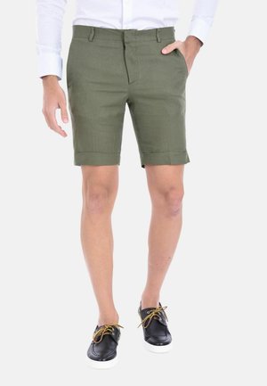 Uomo che indossa pantaloncini verde oliva con orlo arrotolato, camicia bianca a maniche lunghe e scarpe casual nere con lacci marroni, in piedi su sfondo bianco.