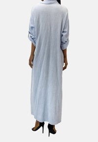 Robe maxi longue bleu clair en tissu texturé. Présente une coupe décontractée, des manches retroussées et un col haut. Portée avec des talons noirs.