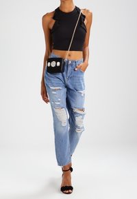 Top corto negro con tirantes acampanados, jeans de mezclilla desgastados en azul claro y bolso cruzado negro con adornos florales. Sandalias de tacón negras.