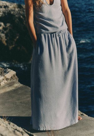 Kobieta ubrana w szarą sukienkę bez rękawów z kieszeniami stoi boso na skalistym brzegu morza w świetle dziennym.