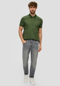Groene polo met kraag, gecombineerd met grijze slim-fit jeans en witte sneakers. De stof lijkt textuur te hebben met een subtiele logo-detail.