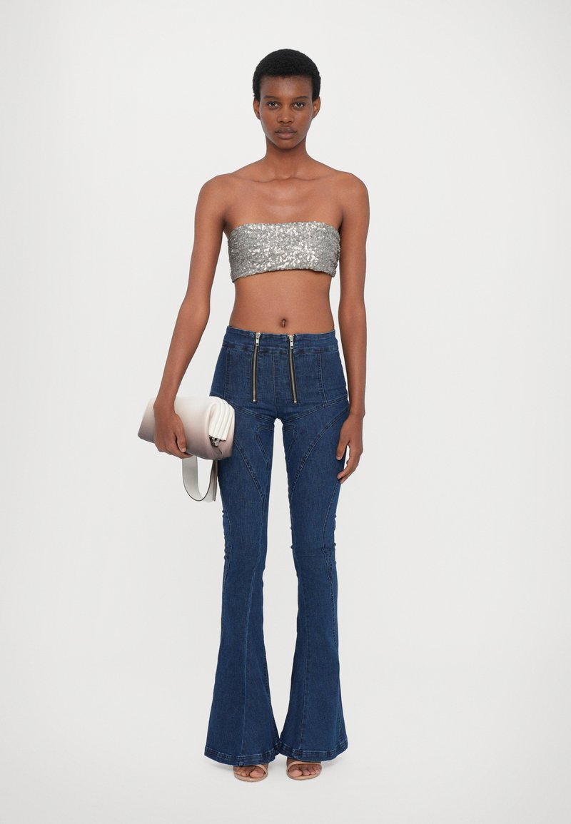 Silverfärgad paljett-topp utan axelband, högmidjade blå utsvängda jeans med dragkedjedetaljer, håller en beige kuvertväska. Släta texturer av tyg.