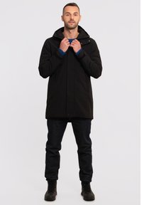 Zwarte lange jas met een capuchon, knopen aan de voorkant en zijzakken. Draagt met donkere jeans en zwarte laarzen. Interieur heeft blauwe accenten.