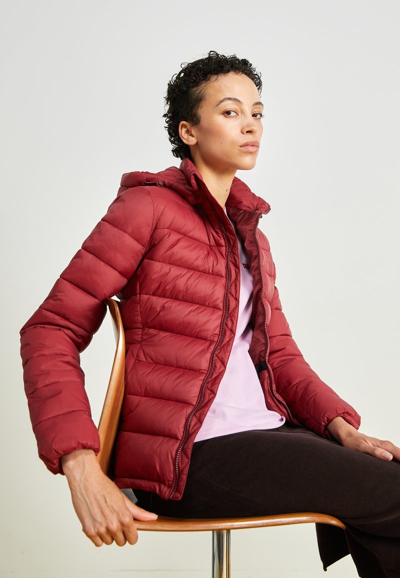 Chaqueta de plumas roja con capucha, patrón acolchado, cremallera frontal y puños elásticos. Lleva puesta una camiseta de color púrpura claro y pantalones negros.