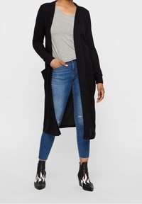 Lång svart cardigan med öppen front, i kombination med en grå V-ringad tröja, slitna blå jeans och svarta ankelstövlar med flammande detaljer.