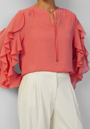 Femme portant un chemisier rose corail à manches à volants et un pantalon blanc plissé taille haute sur fond gris.