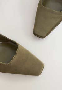 Olijf groene blokhak schoenen met een gladde textielen bovenkant en een vierkant teen ontwerp. De uitgesneden zijkanten onthullen een glanzende binnenvoering.