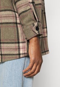 Camisa a cuadros verde y beige con acentos en rosa y negro, que presenta un puño abotonado y una textura suave similar a la lana. Mano descansando sobre un pantalón de mezclilla.