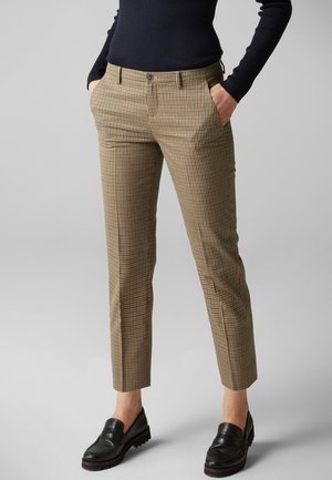 Vrouw die bruine geruite rechte broek draagt, zwarte loafers en een zwarte top met lange mouwen, staand met handen in de zakken.