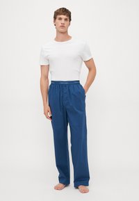 Muškarac stoji bosa, odjeven u bijelu majicu kratkih rukava i široke plave hlače s Calvin Klein oznakom na pojasu, s rukama u džepovima.