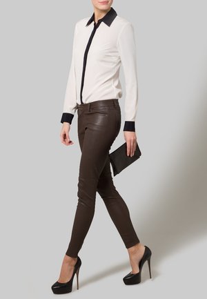 Mujer vestida con blusa blanca con cuello y puños negros, pantalones de cuero marrón, tacones altos negros, sosteniendo un bolso de mano negro, de pie.