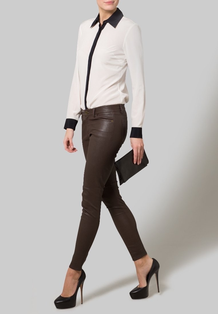 Femme portant un chemisier blanc avec col et poignets noirs, pantalon en cuir marron, talons hauts noirs, tenant une pochette noire, debout.