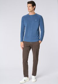 Pullover blu in maglia con scollatura a girocollo, abbinato a pantaloni chinos marroni e sneaker bianche. Il pullover presenta polsini rigati e un logo sul petto.