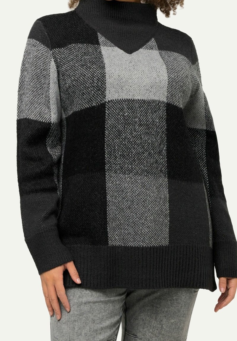 Femme portant un pull à col roulé à grands carreaux noirs et gris avec des poignets et un ourlet côtelés, associé à un pantalon gris clair.