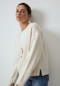 Cardigan beige chiaro realizzato in tessuto a maglia testurizzato, con scollo a V, grandi bottoni frontali e polsini a costine. Abbinato a jeans di denim.