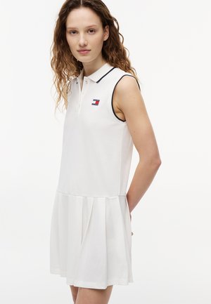Junge Frau mit lockigem Haar, die ein ärmelloses weißes Kleid mit Kragen und plissiertem Rock sowie dem Tommy Jeans-Logo auf der Brust trägt.