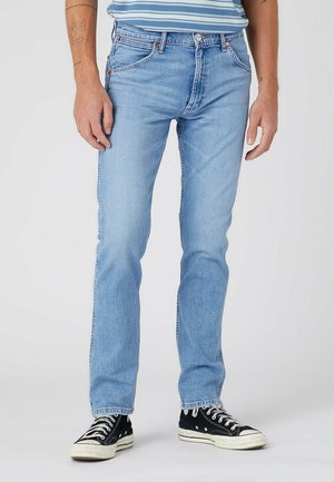 Homme portant un jean slim bleu clair et des baskets montantes noires, debout contre un fond clair uni.