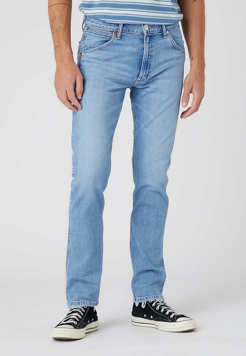 Mann trägt hellblaue Slim-Fit-Jeans und schwarze High-Top-Sneaker, steht vor einem neutralen hellen Hintergrund.