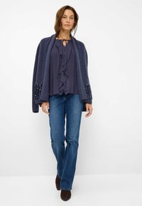 Cardigan in maglia navy con tasche decorate da paillettes, indossato sopra una blusa navy con volant e jeans flare blu, abbinato a stivali marroni.