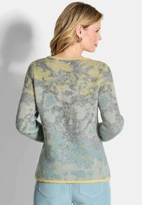 Vrouw met blond haar, die gouden hoepeloren en een fuzzy, multicolor trui in geel, grijs en blauw draagt, met de rug naar ons gekeerd, gecombineerd met lichtblauwe jeans.