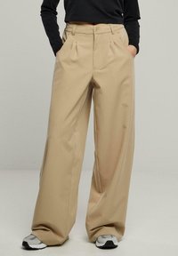Pantalons beige à jambes larges avec des plis, dotés de deux poches latérales et d'une fermeture à bouton, fabriqués dans un tissu lisse et léger.