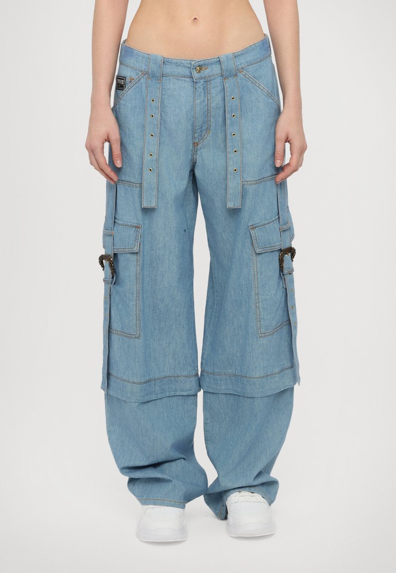 Šviesiai mėlynos džinsinės cargo kelnės su plačiomis kojomis, daugybe kišenių, metaliniais snapeliais ir reguliuojamomis juostos diržais.