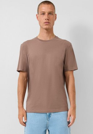 Młody mężczyzna z krótkimi włosami, noszący małe kolczyki kołeczki, prostą brązową koszulkę z krótkim rękawem i jasnoniebieskie dżinsy, stojący na jednolitym tle.