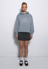 Jasnoniebieska bluza z kapturem z napisem "Local Heroes", zestawiona z czarną plisowaną mini spódnicą. Białe skarpetki do kostek i brązowe buty Mary Jane.