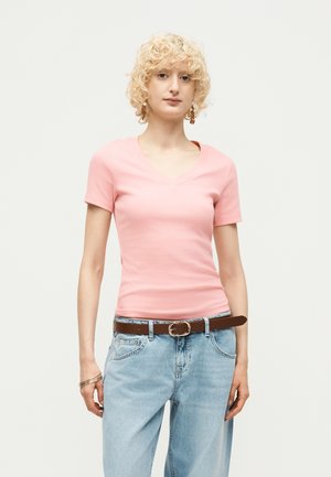 Jeune personne aux cheveux blonds bouclés portant un t-shirt rose à manches courtes et col en V, un jean bleu clair, une ceinture marron, ainsi que des boucles d'oreilles et bracelets en or.