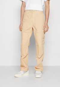 Beige cargo pants made of lightweight fabric, featuring side pockets, stitched seams, and a relaxed fit. Worn with white sneakers.