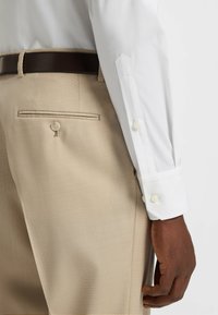 Chemise blanche à manches longues avec des poignets boutonnés, assortie à un pantalon beige avec une poche arrière et un détail de bouton, texture de tissu lisse.