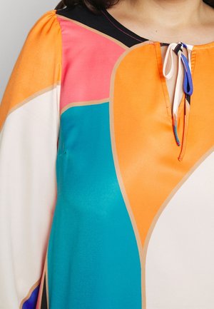 Blouse à manches longues avec un motif géométrique coloré en orange, bleu canard, rose, noir et blanc, avec un décolleté en trou de serrure noué par un cordon bouclé.