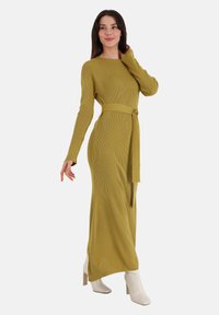 Abito lungo a coste in verde oliva con maniche lunghe e cintura annodata in vita. Abbinato a stivaletti bianchi alla caviglia, che mostrano una silhouette elegante.