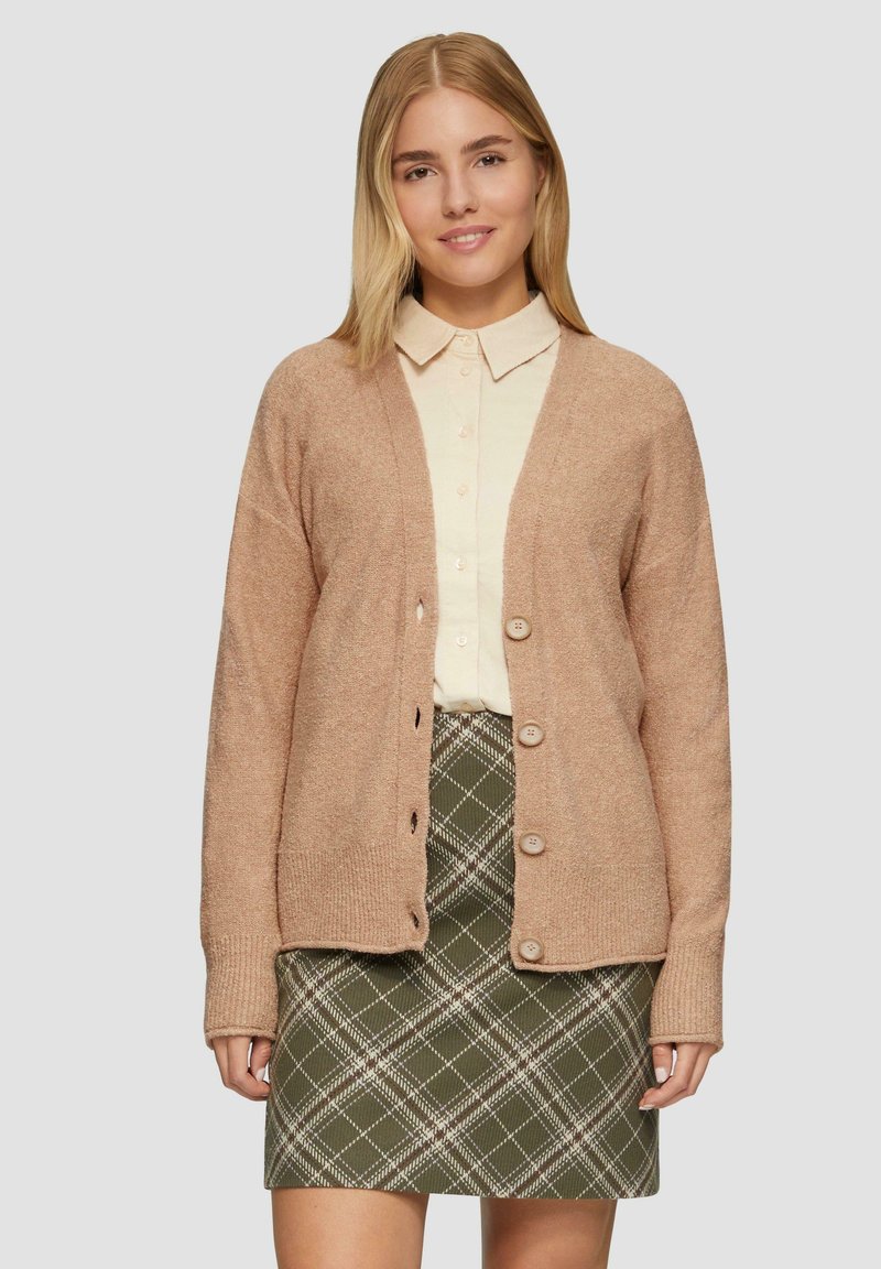 Cardigan lavorato a maglia rosa chiaro con scollo a V, maniche lunghe e cinque bottoni. Abbinato a una camicia color crema e una gonna a quadri verde.