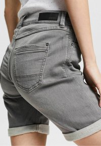 Zbliżenie na osobę noszącą podwinięte szare dżinsowe szorty, z widoczną tylną kieszenią oraz metką marki Esprit na pasku.